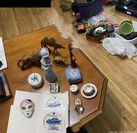 Full view of lot with wood carvings, porcelain items, patches, mask, and glass shoe on a wooden table in a room with hardwood floor