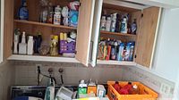 Wide shot of multiple shelves inside wooden cupboard with assorted cleaning supplies arranged and some on sink area below.