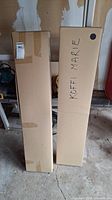 Two sealed cardboard boxes labeled 'KOFFI MARIE' and another with a sticker, presumably containing shelving units, placed on a concrete floor in a garage.