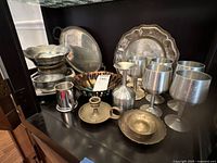 Full view of all metalware items arranged on a shelf including goblets, trays, bowls, and candlestick holder with visible tarnishing.