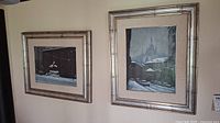 Two framed snow-themed urban scene paintings side by side on wall with silver-tone frames and beige mats.