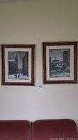 Two framed signed snow-themed paintings hanging on wall above couch, depicting winter urban scenes.