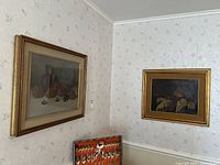 Two framed paintings hanging on wall, photographed from the corner showing the setting and sizes.