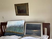 Three pieces of original art displayed on a chair: two framed and one canvas board showing landscapes and nature scenes, with frames and canvas visible