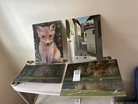 Four paintings displayed on a white surface showing a fox, a village street, and two landscape scenes.