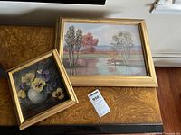 Two framed paintings on wooden table: floral still life and landscape.
