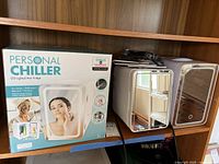 One new Personal Chiller LED Lighted Mini Fridge in original packaging and two used units with power cords on a wooden shelf.