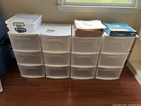 Four sets of white translucent plastic storage drawers each with three drawers arranged side by side; several packs of paper file folders on top
