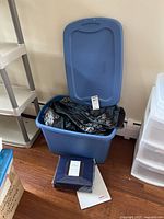 Blue plastic storage bin open with bedding inside and queen sheet set box on floor