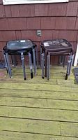Two stacks of black framed side tables with tempered glass tops, each stack containing 3 tables. The tables appear to be used and located outdoors on a wooden deck.
