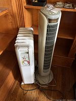 Side-by-side view of Lakewood heater and Aloha Breeze tower fan on wood floor near wooden shelves.