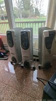 Three Pelonis electric radiator space heaters with off-white ribbed oil-filled radiators on wheels. Positioned indoor near window on tiled floor. Power cords visible. Moderate surface wear.