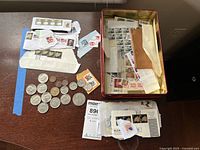 US coins and sheets/strips of US postage stamps on envelopes and loose pieces of paper, including pennies, nickels, dimes, quarters, half dollars, and dollar coin.
