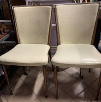 Front view of two vintage MCM dining chairs with cream vinyl seats and backs and teak frames.