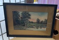 Framed watercolour landscape painting with trees, grassy field, signed in lower right corner and behind glass in a dark wooden frame with yellow mat.
