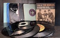 Photo showing a lot of 8 Beatles vinyl 45 RPM records including one jacket for "Four by The Beatles." Records have visible wear and some plain sleeves.