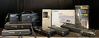 Wide shot of vintage voice recording devices including microcassette recorders and cassette players from Sony, Sanyo, and others, arranged on a flat surface against black background.