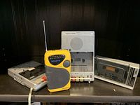 Photo showing a yellow AM/FM radio and three vintage cassette tape recorders/decks on a shelf.