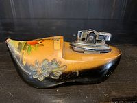 Novelty vintage wooden clog shaped table top lighter with painted floral and scenic designs and attached metal lighter mechanism.