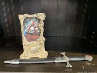 Front view showing a metal dagger sheathed, placed horizontally in front of a decorative card stand featuring a movie card.