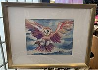Framed watercolour painting showing an owl in flight against a cloudy sky. The owl has spread wings with shades of purple and brown, mounted in a light wood frame with matting.