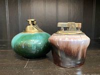 Photo shows two vintage pottery table top lighters. The green one is marked BMP (Blue Mountain Pottery); the brown and cream one is unmarked. Both have metal lighter tops with visible wear.