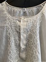 Close-up photo of the neckline with detailed hand embroidery showing floral and geometric patterns in white thread on white silk fabric, with buttons.