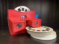 Vintage red 3D View-Master toy viewer with multiple slide reels stacked beside it, showing slots for inserting reels and dual lenses for 3D viewing.