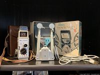 Image showing vintage Bell & Howell film camera with its original brown leather carrying case next to a Baia film reviewer with two reels, original box, and power cord.