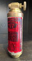 Photo showing the front side of the vintage brass Presto B mini fire extinguisher with worn red label and brass top valve