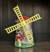 Front view of the vintage tin windmill toy showing colorful illustrations of a cow, flowers, and the yellow plastic blades.