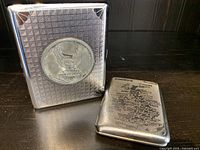 Front view showing both vintage metal cigarette/card cases: Harley Davidson case with eagle emblem and geometric texture, and British Isles case with engraved map of locations.
