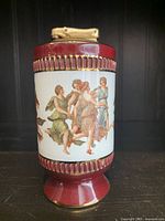 Side view of vintage table lighter showing classical Renaissance-style painted figures in flowing garments, on a deep red base with gold trim.