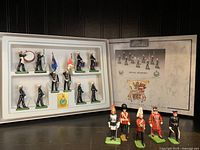 Photo showing one open box of ten mint condition metal British Royal Marines toy soldiers in dark uniforms with white pith helmets and musical instruments or flags, along with five loose brightly painted metal British toy soldier figures in a variety of uniforms and poses on green bases.