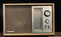 Front view of vintage Panasonic transistor radio with wood laminate casing, fabric speaker grille, analog AM tuning scale, and control knobs.