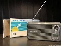 Vintage Realistic transistor radio with extended telescopic antenna, frequency dial, and AM/FM switch. Original box and insert shown behind it.
