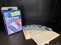 Photo of the vintage Yahtzee travel game box alongside six printed score sheets spread out.
