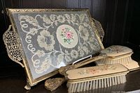 Photo showing four-piece vintage ladies dresser set with ornate metal and embroidered tray, hand mirror, and two floral painted hair brushes.
