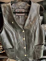 Photo of black leather motorcycle vest laid flat showing front snap button closure and braided leather trim.
