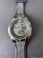 Front view of the digital wristwatch showing the time 3:05 with day, date, and chronograph display. Black textured rubber strap and stainless steel case with labeled bezel buttons.