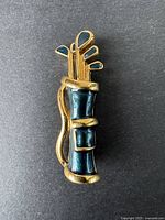 Brooch in gold-tone metal shaped as a golf bag with clubs, showing black enamel detailing and gold bands.
