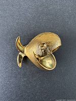 Photo of gold-tone whale-shaped brooch on black background, showing front view, medium-sized with textured surface.