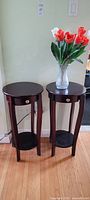 Two dark wood round plant stands or occasional tables side by side on a wood floor, one holding a glass vase with silk flowers