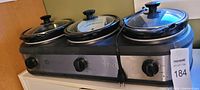 Front view of three 3-quart slow cookers and one warming unit on an integrated electrical base, showing stainless steel, black knobs, and glass lids with handles.