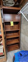 Full view of the tall, brown wooden bookcase with five shelves, placed in a corner near cinder block walls.