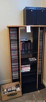 Front view of tall CD cabinet showing CDs in side racks, some cassettes and DVDs on shelves, and cardboard box with cassettes at base.
