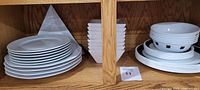 Wooden cabinet shelf with stacked white round plates, white diamond-shaped dish stack, and white bowls with black square pattern.