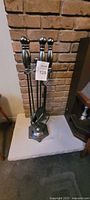 Fireplace tool set with poker, brush, and shovel on stand in front of brick fireplace base.