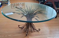 Table shown at slight angle on hardwood floor, displaying round glass top and full wheat-themed base.