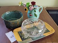 Green pottery bowl, light green ceramic pitcher with porcelain flowers, clear medium crystal bowl on yellow plastic tray, all on wooden table